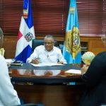 Director General de Migración firmando acuerdo junto al Director de Planificación y su asistente, sentados en el despacho con persianas de madera y banderas de fondo.