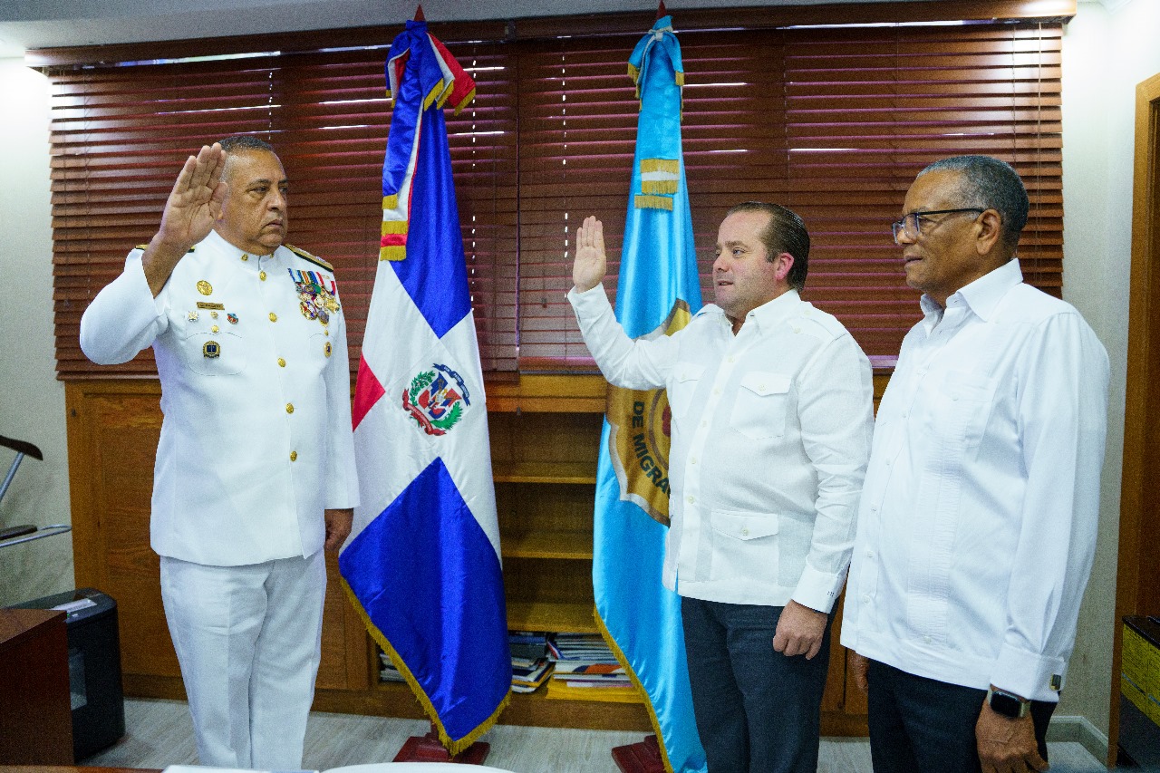 Juramentación de nuevo director general de migración en el despacho levantando la mano con Ignacio Paliza y el anterior Director, persianas de madera y bandera de fondo.