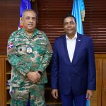 Director General in camouflage uniform next to Victor de Aza President of the Dominican Municipal League in blue suit pose in front of flags and a background of wooden shutters in the DGM office.