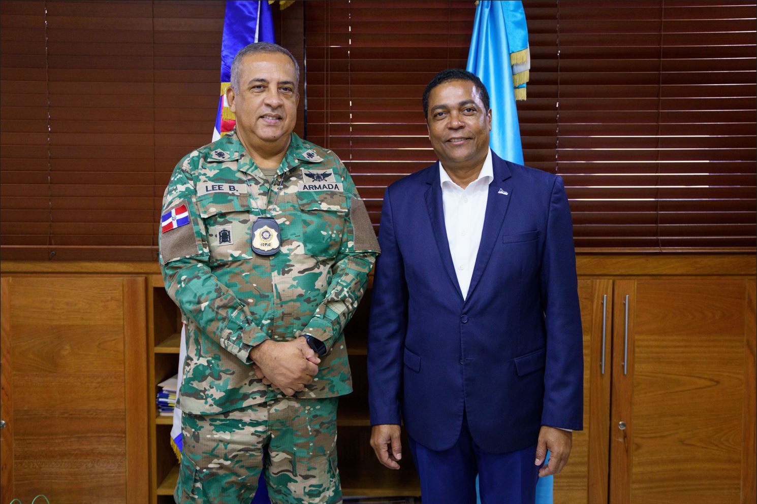 Director General in camouflage uniform next to Victor de Aza President of the Dominican Municipal League in blue suit pose in front of flags and a background of wooden shutters in the DGM office.