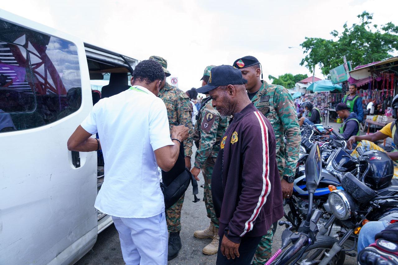 Oficiales DGM revisando extranjero en operativo frente a minibús blanco y parada de motoristas.