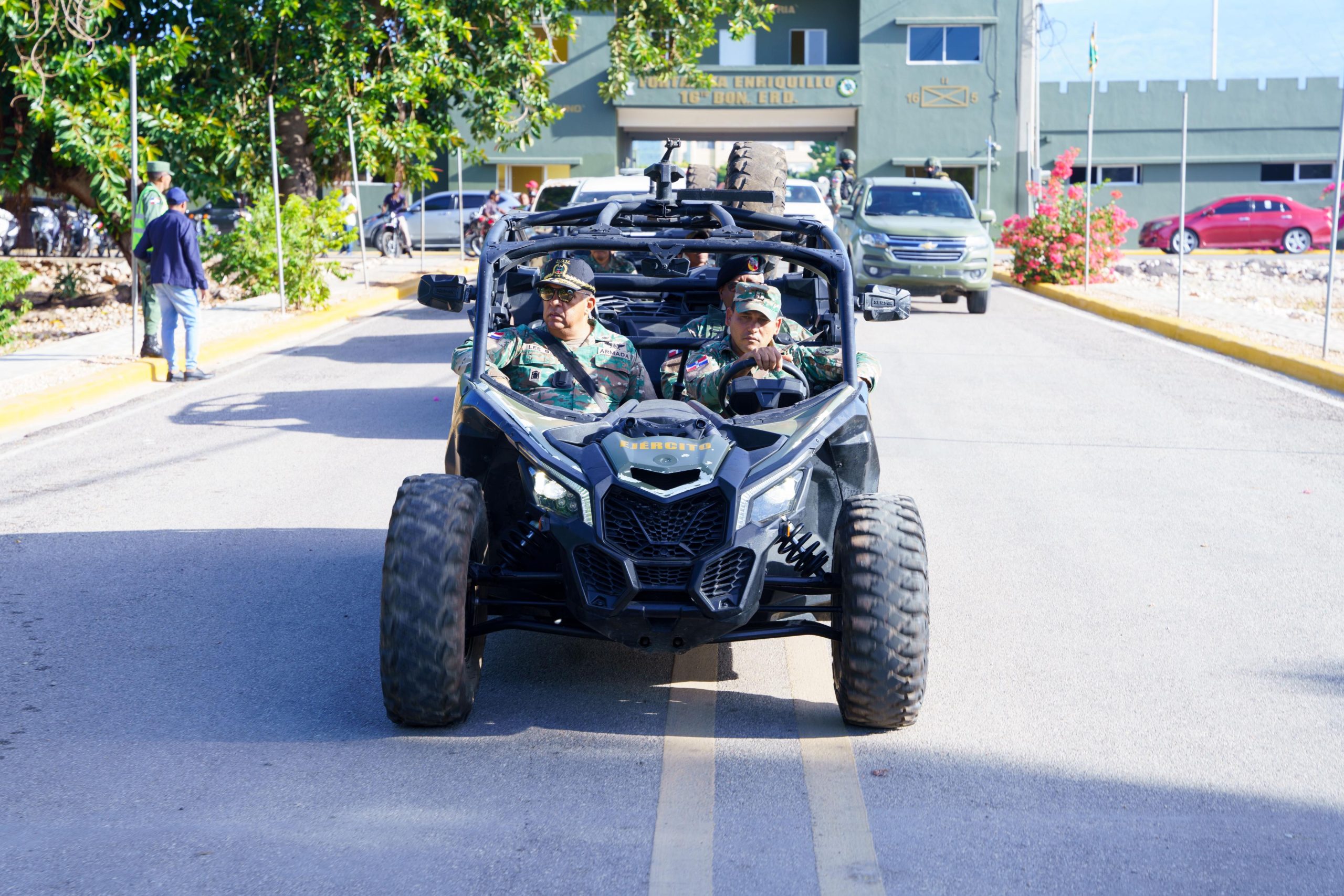 Director General together with other officers in camouflage uniform in a military raptor buggy in front of Fortaleza Enriquillo, with vehicles and people in the background.