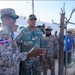 Dominican military personnel along with the Director General in camouflage uniforms interact with civilians in front of a metal fence in an open-air setting at the border under clear skies.