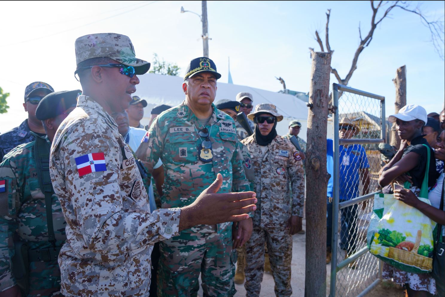 Dominican military personnel along with the Director General in camouflage uniforms interact with civilians in front of a metal fence in an open-air setting at the border under clear skies.