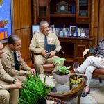 Reunión de Director General con procuradora general de la República Dominicana con otros directores departamentales de la DGM sentados alrededor de una mesa decorada con plantas, libros y una gorra militar.