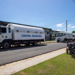 Camiones blancos de la Dirección General de Migración, identificados como "Control Migratorio", estacionados frente a un edificio bajo un cielo despejado, con motocicletas aparcadas al lado derecho..