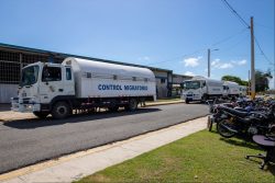 Camiones blancos de la Dirección General de Migración, identificados como "Control Migratorio", estacionados frente a un edificio bajo un cielo despejado, con motocicletas aparcadas al lado derecho..