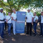 Un grupo de personas con camisetas blancas y guantes sostiene plantas frente a un cartel que dice "Jornada de Reforestación", en un entorno al aire libre con árboles al fondo.