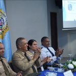 Director General among other authorities sitting at a table in the Juan Pablo Duarte room of the DGM applauding and smiling.