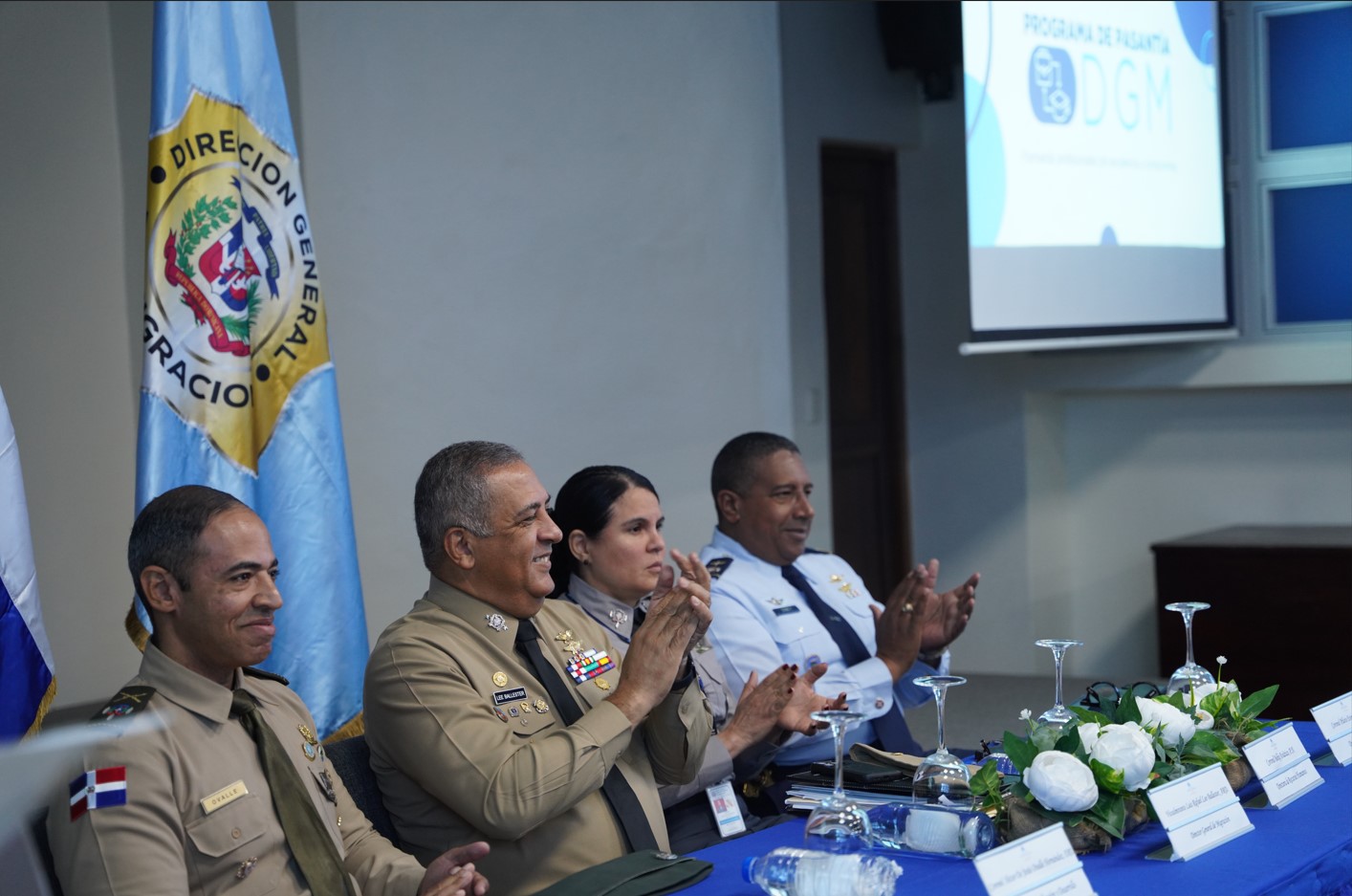 Director General among other authorities sitting at a table in the Juan Pablo Duarte room of the DGM applauding and smiling.