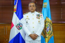 Director General parado alate de la bandera dominicana y la bandera de la DGM posando con brazos abajo y con las manos una arriba de la otra.