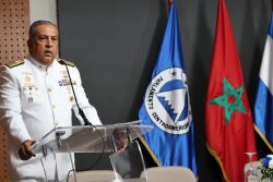 Director de pie detrás de podium, con banderas de diferentes nacionalidades al lado, dando conferencia